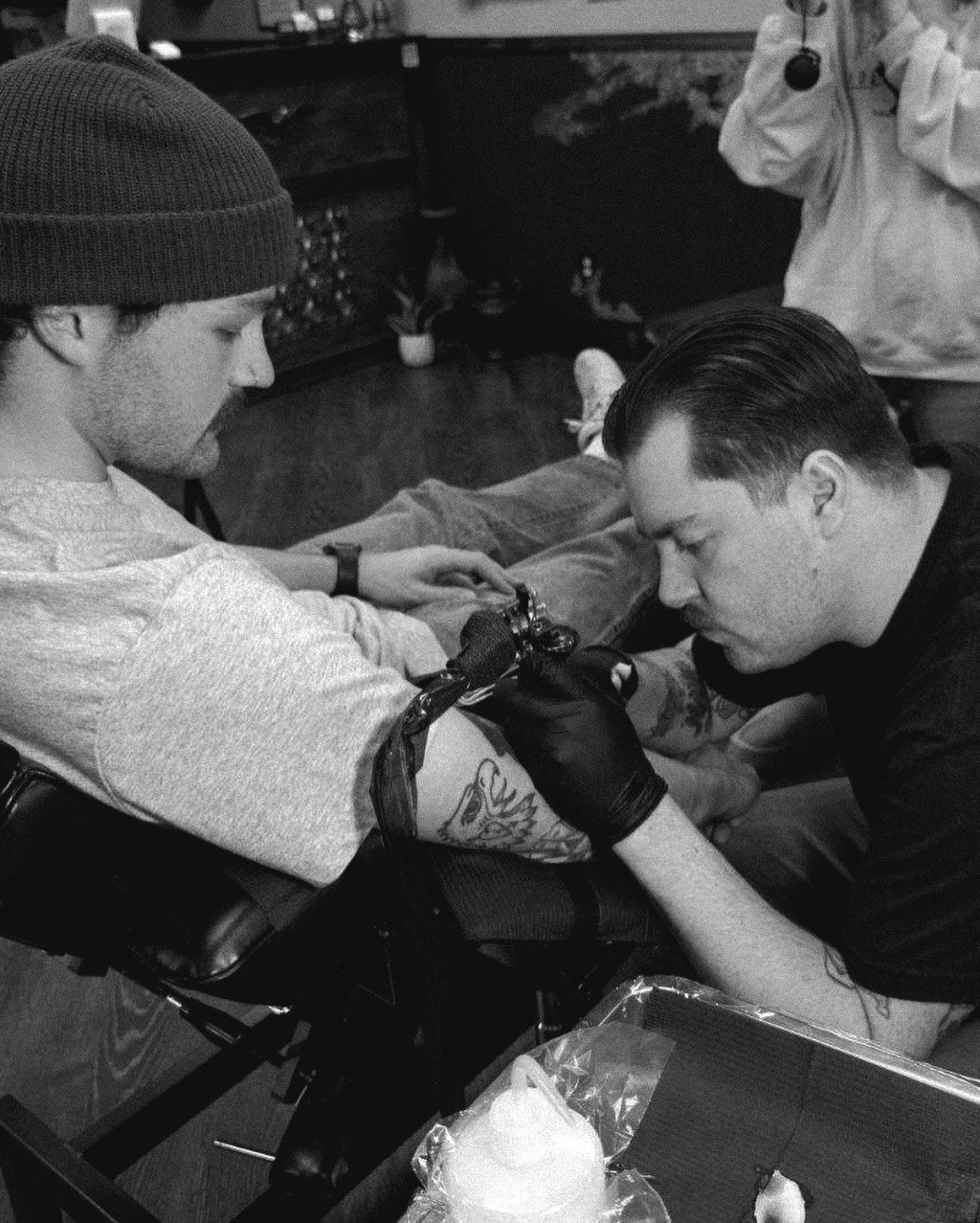 "Apprentice tattoo artist Dru at Lady Liberty Tattoo working on a client’s arm. Black and white photo captures the focus and precision of tattooing in a professional studio setting."
